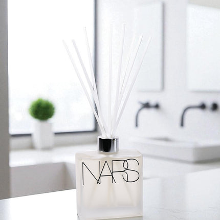 A frosted glass reed diffuser with black text and white reed sticks sits on a white marble countertop in a modern bathroom, with white subway tile backsplash, black fixtures, and a small green plant visible in the background