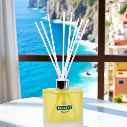 A frosted glass reed diffuser with tinted yellow fragrance; branding and white reed sticks sits on a white round table by a window, overlooking a stunning Mediterranean coastal view with turquoise waters, dramatic cliffs, and colorful hillside buildings