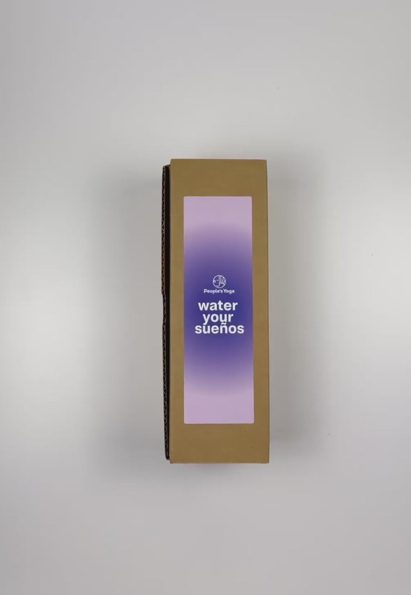 An unboxing video of the 16 oz Prayer Candle from Custom Candles LA filled with purple wax that comes in a Kraft mailer box with a diffused purple sticker label of logo artwork. The candle is printed with a white "People's Yoga" logo and underneath it reads "water your suenos" in white. Behind the letters is a blue circle and fades into a lighter purple color. You can see the purple wax through the glass.