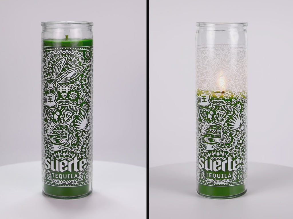 Two 16 oz Prayer Candles from Custom Candles LA filled with green wax in a tall glass container spinning side by side. The candles are printed a logo at the bottom that reads "Suerte Tequila" in white. The logo is surrounded by an ornate pattern that makes the shape of a rabbit. The pattern includes diamonds, dots, flowers, and other line artwork. The green wax shows through the artwork.