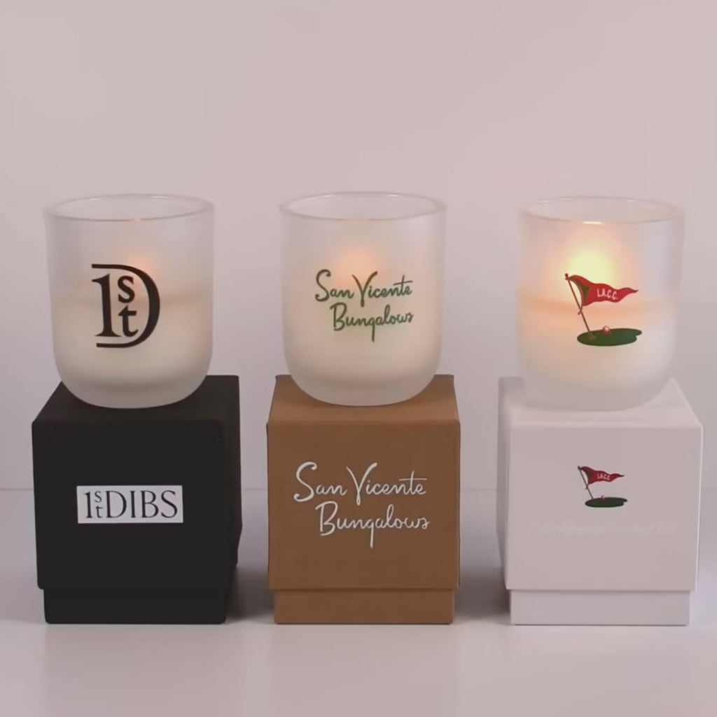 Three lit 12 oz Luxe Custom Candles in the color “Cashmere” which is a diffused white from Custom Candles LA. The 1st candle has a black "1st Dibs" logo printed on the glass and sits on a black box that says "1st Dibs" printed in white. The 2nd candle has a "San Vicente Bungalows" logo printed in green and sits on top of a Kraft box with the same logo printed on it in white. The 3rd candle has the "L.A.C.C" logo printed on the glass in multicolor, and sits on a white box with the same logo printed. 