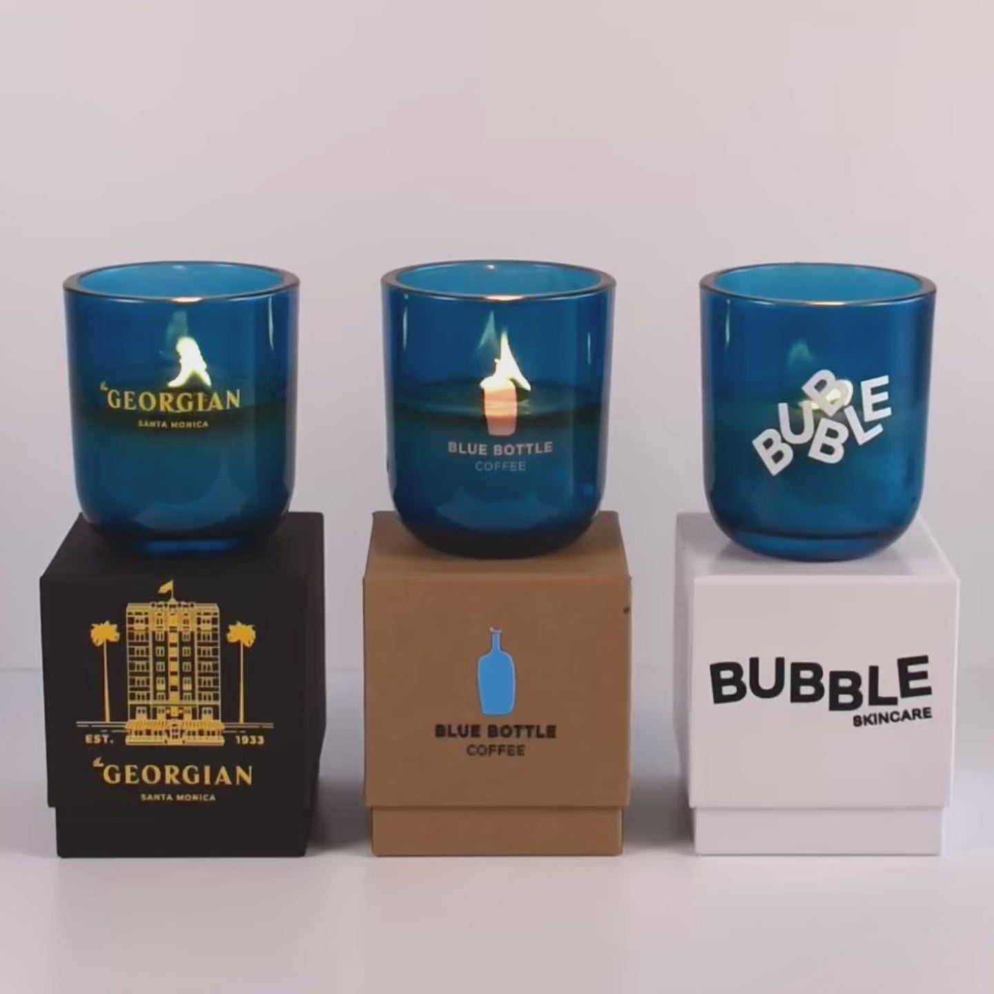 Load video: Three lit 12 oz Luxe Custom Candles in the color “Oceania” which is a splashy teal from Custom Candles LA. The 1st candle has a yellow &quot;Georgian&quot; logo printed on the glass and sits on a black box that also has the printed yellow logo. The 2nd candle has an etched &quot;Blue Bottle Coffee&quot; logo on the glass and sits on top of a Kraft box with the logo printed on it in blue and black. The third candle has the logo &quot;BUBBLE&quot; printed on the glass in white, and sits on a white box with the logo printed in black.