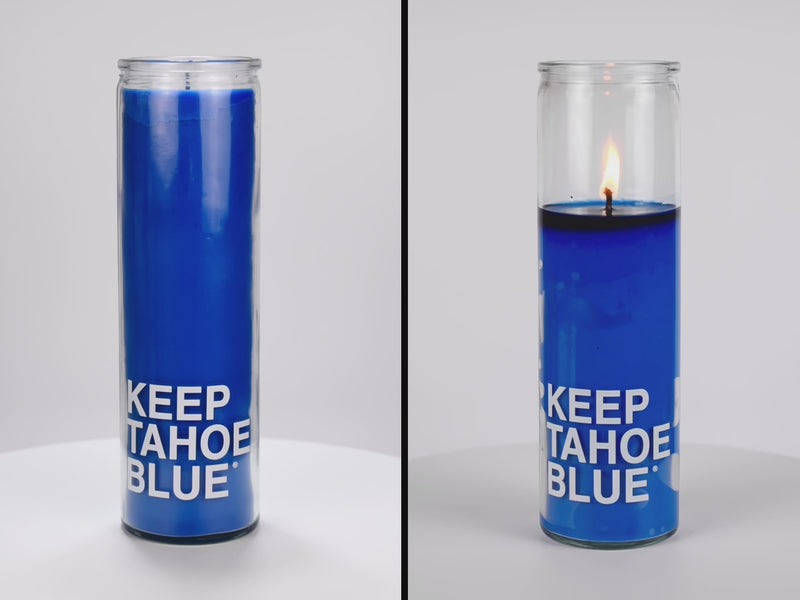 Two 16 oz Prayer Candles from Custom Candles LA filled with blue wax in a tall glass container spinning side by side. The left candle is unlit, and the right candle is lit and burned halfway down. The candles are printed with the words "KEEP TAHOE BLUE" in white letters across the bottom of the candle. 