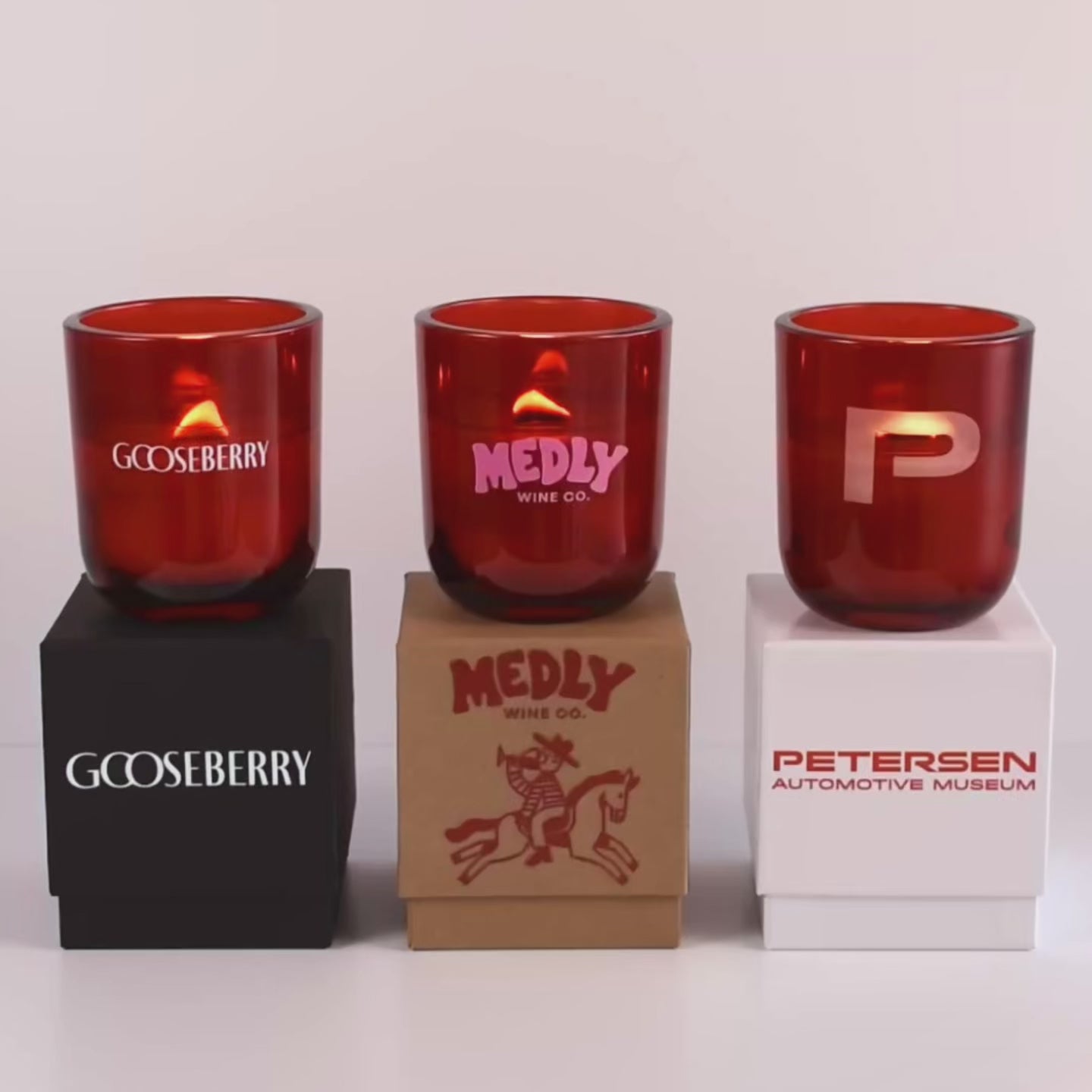 Load video: Three lit 12 oz Luxe Custom Candles in the color “Blood Red” which is a vivid rouge from Custom Candles LA. The 1st candle has the &quot;Gooseberry&quot; logo printed on the glass in white and sits on a black box that has the same logo printed on it. The 2nd candle has a pink &quot;Medly Wine Co.&quot; logo printed on the glass and sits on top of a Kraft box with the logo printed in red. The 3rd candle has a &quot;P&quot; etched on the glass, and sits on a white box with the logo &quot;Petersen Automotive Museum&quot; printed in red.