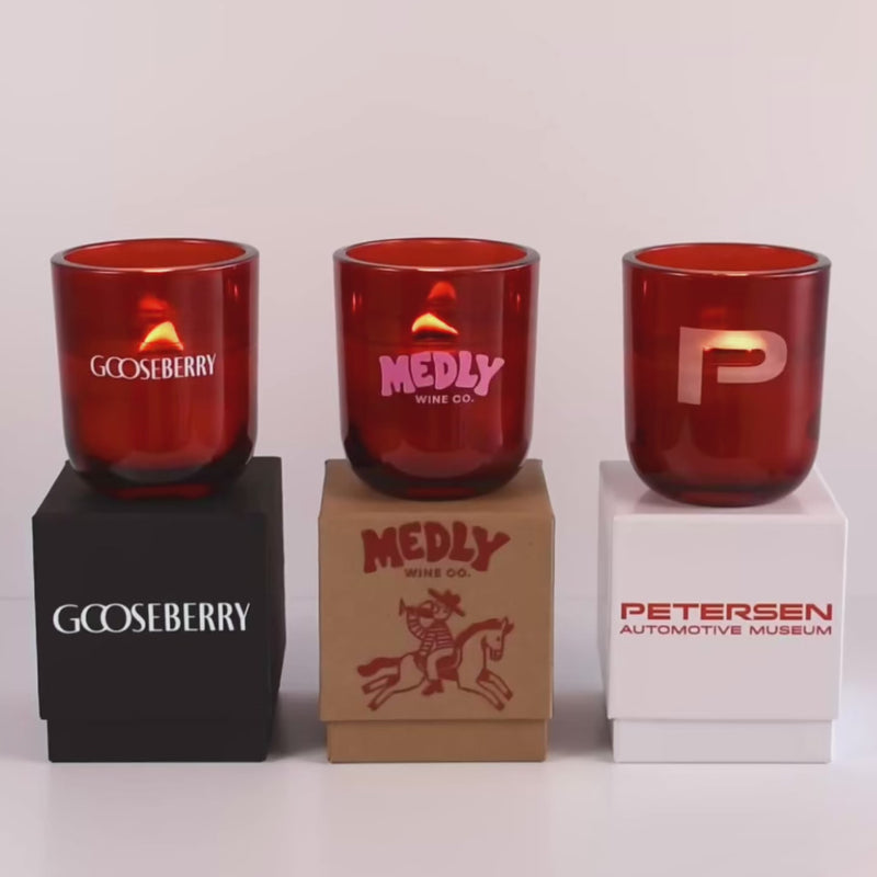 Three lit 12 oz Luxe Custom Candles in the color “Blood Red” which is a vivid rouge from Custom Candles LA. The 1st candle has the "Gooseberry" logo printed on the glass in white and sits on a black box that has the same logo printed on it. The 2nd candle has a pink "Medly Wine Co." logo printed on the glass and sits on top of a Kraft box with the logo printed in red. The 3rd candle has a "P" etched on the glass, and sits on a white box with the logo "Petersen Automotive Museum" printed in red.