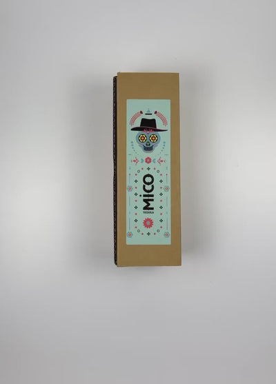 An unboxing video of the 16 oz Prayer Candle from Custom Candles LA filled with yellow wax that comes in a Kraft mailer box with a teal sticker label. The candle is printed with a colorful image of a calavera surrounded by patterns in various colors and shapes including flowers, dots, and diamonds. In the middle of the candle there is a vertical logo printed in black that reads "MICO TEQUILA". The yellow wax shows through the artwork