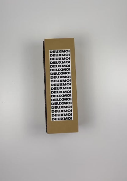 An unboxing video of the 16 oz Prayer Candle from Custom Candles LA filled with gray wax that comes in a Kraft mailer box with a black and white sticker label of the logo. The candle is printed with a logo that reads "DEUXMOI" in bold black letters that are stacked vertically down the front of the glass.