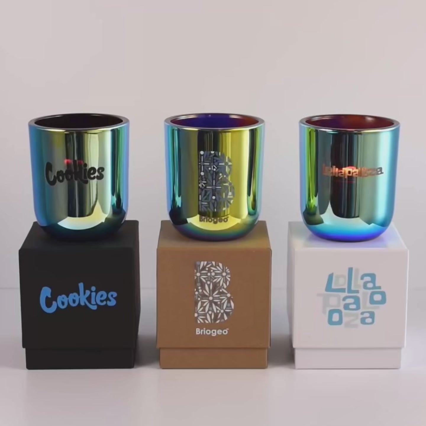 Three lit 12 oz Luxe Custom Candles in the color “Euphoria” which is a metallic blue from Custom Candles LA. The first candle has a black "Cookies" logo printed on the glass and sits on a black box that has the same logo in blue. The second candle has a multicolor "Briogeo" logo on the glass and sits on top of a Kraft box with the same logo printed on it. The third candle has the "Lollapalooza" logo etched on the glass, and sits on a white box with the logo printed in different blues.
