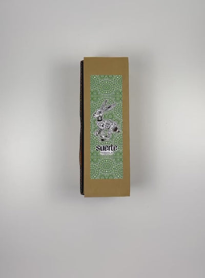 An unboxing video of the 16 oz Prayer Candle from Custom Candles LA filled with green wax that comes in a Kraft mailer box with a green sticker label of the logo artwork. The candle is printed a logo at the bottom that reads "Suerte Tequila" in white. The logo is surrounded by an ornate pattern that makes the shape of a rabbit. The pattern includes diamonds, dots, flowers, and other line artwork. The green wax shows through the artwork.