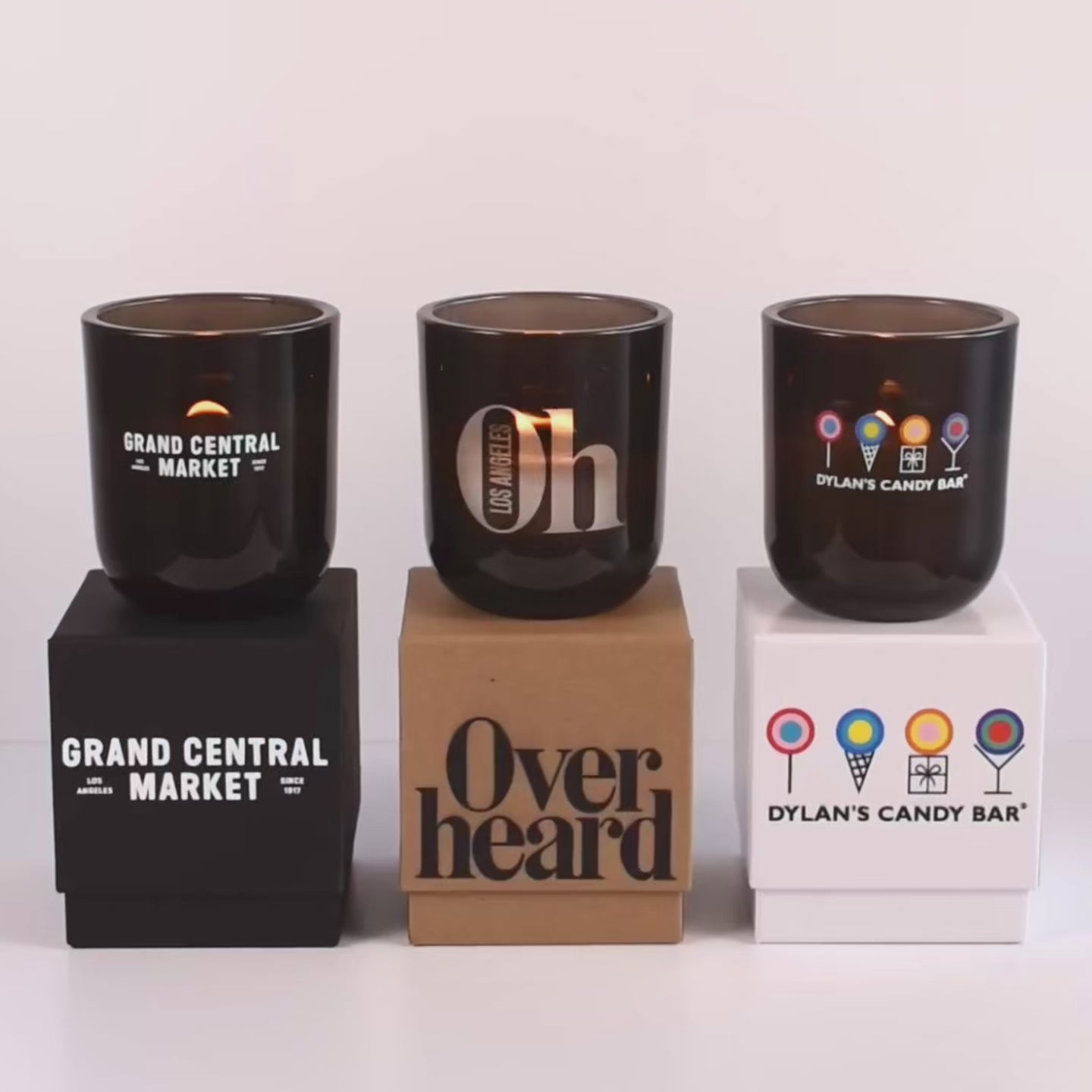 Load video: Three lit 12 oz Luxe Custom Candles in the color “Nightfall” which is a smoke grey from Custom Candles LA. The 1st candle has the &quot;Grand Central Market&quot; logo printed on the glass in white and sits on a black box that has the same logo printed on it. The 2nd candle has a &quot;Oh Los Angeles&quot; logo etched on the glass and sits on top of a Kraft box with the logo &quot;Overheard&quot; printed in black. The 3rd candle has &quot;Dylan&#39;s Candy Bar&quot; logo printed in multicolor and sits on a white box with the same logo printed on it. 