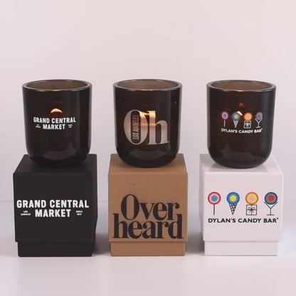 Three lit 12 oz Luxe Custom Candles in the color “Nightfall” which is a smoke grey from Custom Candles LA. The 1st candle has the "Grand Central Market" logo printed on the glass in white and sits on a black box that has the same logo printed on it. The 2nd candle has a "Oh Los Angeles" logo etched on the glass and sits on top of a Kraft box with the logo "Overheard" printed in black. The 3rd candle has "Dylan's Candy Bar" logo printed in multicolor and sits on a white box with the same logo printed on it. 