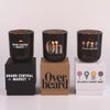 Three lit 12 oz Luxe Custom Candles in the color “Nightfall” which is a smoke grey from Custom Candles LA. The 1st candle has the "Grand Central Market" logo printed on the glass in white and sits on a black box that has the same logo printed on it. The 2nd candle has a "Oh Los Angeles" logo etched on the glass and sits on top of a Kraft box with the logo "Overheard" printed in black. The 3rd candle has "Dylan's Candy Bar" logo printed in multicolor and sits on a white box with the same logo printed on it. 