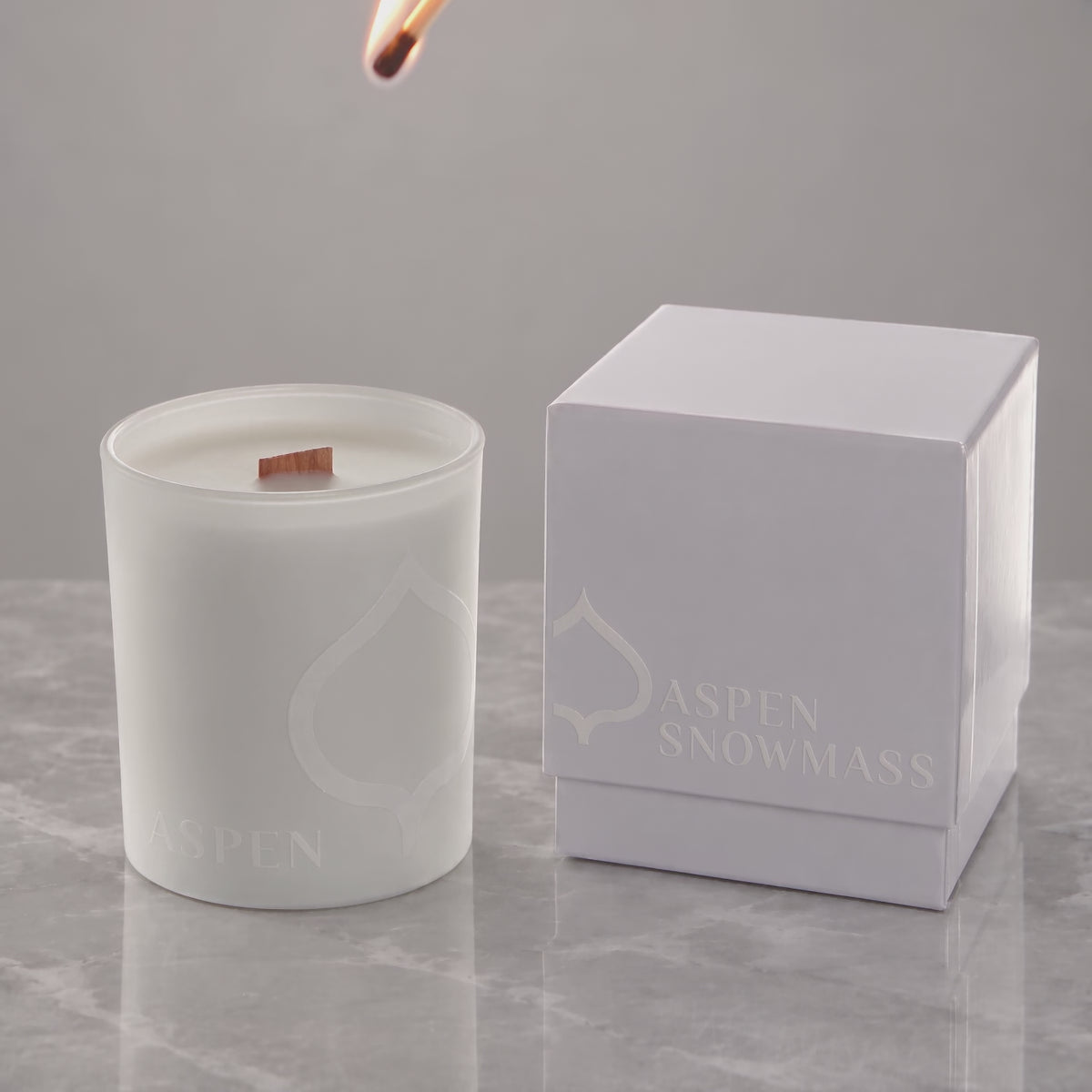 White 14 oz. printed luxury candle lit, with a tone-on-tone Aspen Snowmass logo glowing beside its white gift box