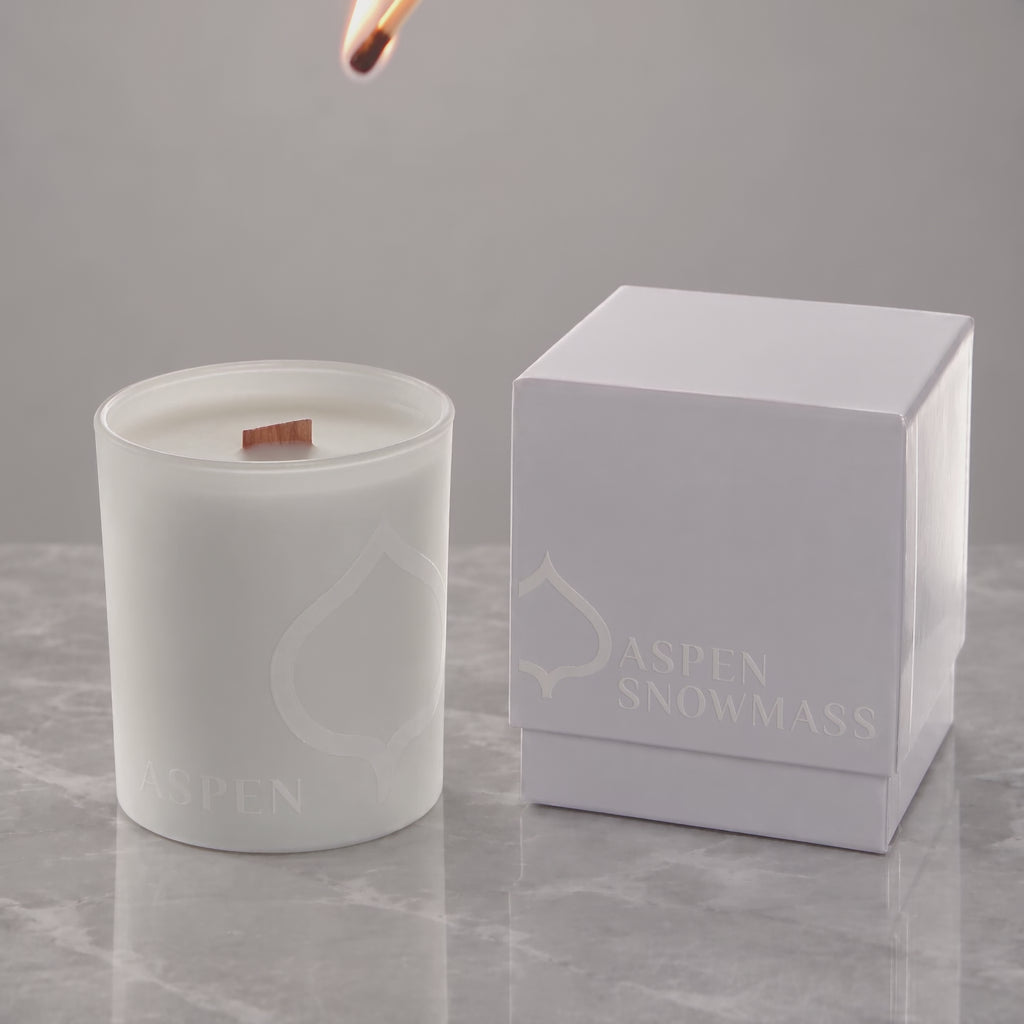 White 14 oz. printed luxury candle lit, with a tone-on-tone Aspen Snowmass logo glowing beside its white gift box
