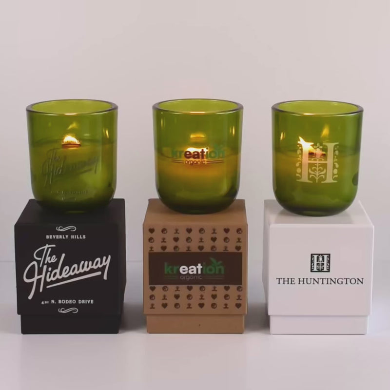 Three lit 12 oz Luxe Custom Candles in the color “Grassland” which is a verdant green from Custom Candles LA. The 1st candle has a green "The Hideaway" logo printed on the glass and sits on a black box that has the logo printed in white. The second candle has a green and brown "kreation" logo on the glass and sits on top of a Kraft box with the multicolor logo printed on it. The third candle has an "H" logo etched on the glass, and sits on a white box with the logo "The Huntington" printed on it in black.
