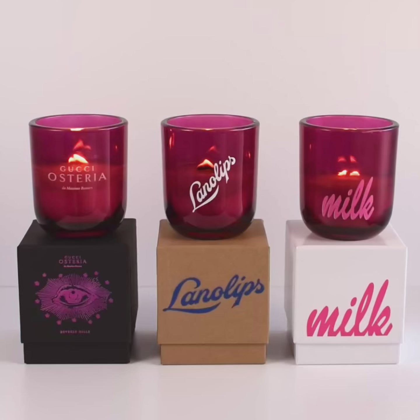 Load video: Three lit 12 oz Luxe Custom Candles in the color “Cherry Kiss” which is a juicy magenta from Custom Candles LA. The 1st candle has the &quot;Gucci Osteria&quot; logo etched on the glass and sits on a black box that has the logo printed in hot pink. The 2nd candle has a white &quot;Lanolips&quot; logo printed the glass and sits on top of a Kraft box with the same logo printed on it in blue. The 3rd candle has the &quot;milk&quot; logo printed on the glass in pink, and sits on a white box with the same logo printed on it in pink.