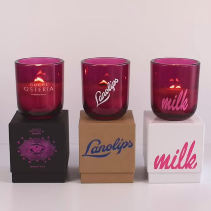 Three lit 12 oz Luxe Custom Candles in the color “Cherry Kiss” which is a juicy magenta from Custom Candles LA. The 1st candle has the "Gucci Osteria" logo etched on the glass and sits on a black box that has the logo printed in hot pink. The 2nd candle has a white "Lanolips" logo printed the glass and sits on top of a Kraft box with the same logo printed on it in blue. The 3rd candle has the "milk" logo printed on the glass in pink, and sits on a white box with the same logo printed on it in pink.