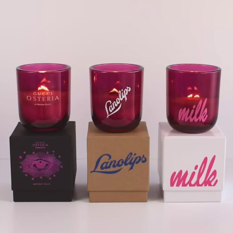 Three lit 12 oz Luxe Custom Candles in the color “Cherry Kiss” which is a juicy magenta from Custom Candles LA. The 1st candle has the "Gucci Osteria" logo etched on the glass and sits on a black box that has the logo printed in hot pink. The 2nd candle has a white "Lanolips" logo printed the glass and sits on top of a Kraft box with the same logo printed on it in blue. The 3rd candle has the "milk" logo printed on the glass in pink, and sits on a white box with the same logo printed on it in pink.