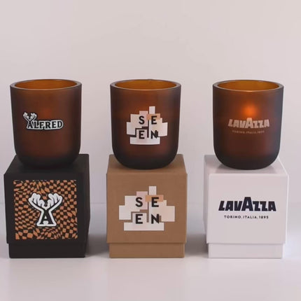 Three lit 12 oz Luxe Custom Candles in the color “Cacao” which is an amber brown from Custom Candles LA. The 1st candle has a black and white "ALFRED" logo printed on it and sits on a black box that also has a checkmark pattern and "A" logo. The 2nd candle has a printed "SEEN" logo on the glass and sits on top of a Kraft box with the same logo. The 3rd candle has the logo "LAVAZZA" etched on the glass, and sits on a white box with the same logo printed in dark blue.