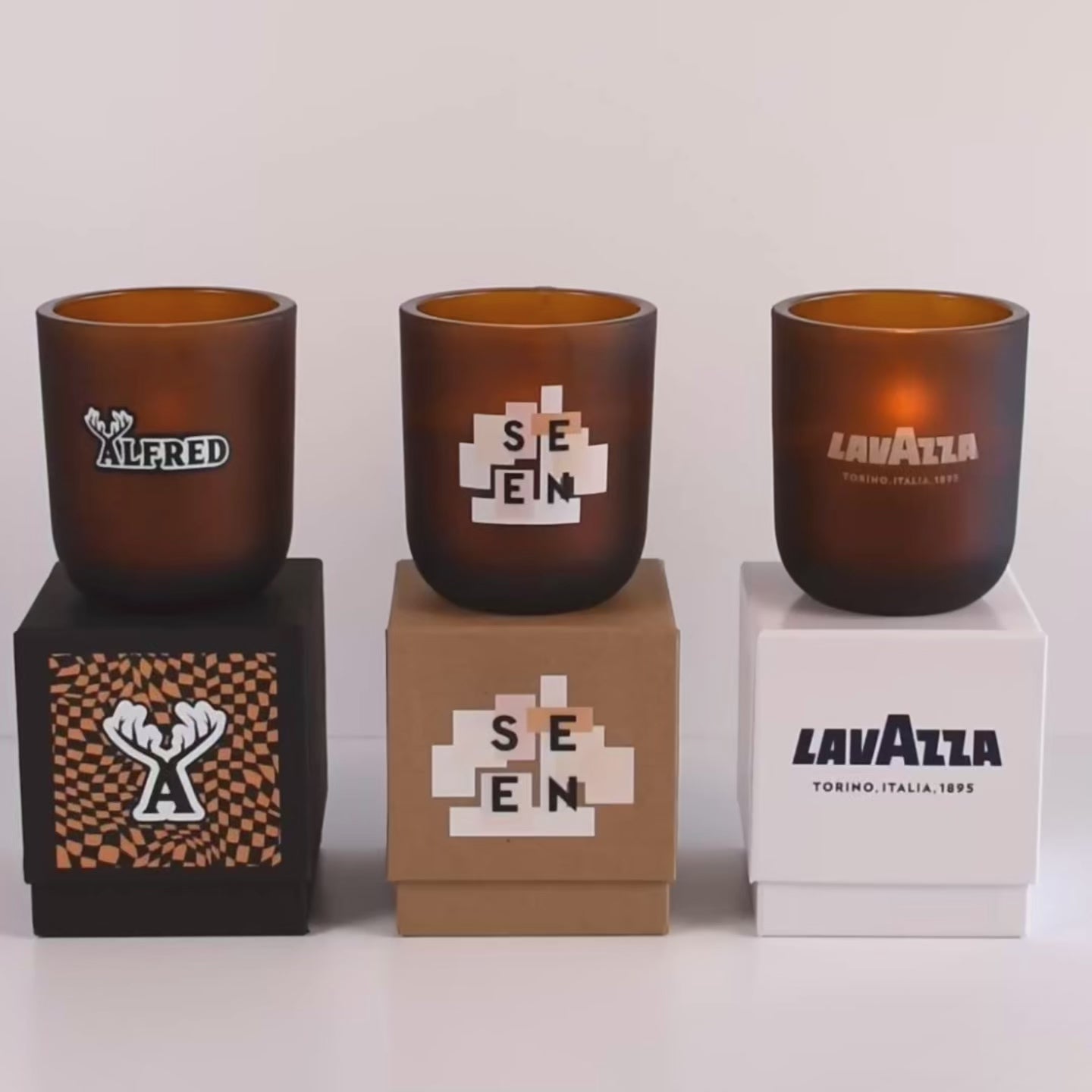 Load video: Three lit 12 oz Luxe Custom Candles in the color “Cacao” which is an amber brown from Custom Candles LA. The 1st candle has a black and white &quot;ALFRED&quot; logo printed on it and sits on a black box that also has a checkmark pattern and &quot;A&quot; logo. The 2nd candle has a printed &quot;SEEN&quot; logo on the glass and sits on top of a Kraft box with the same logo. The 3rd candle has the logo &quot;LAVAZZA&quot; etched on the glass, and sits on a white box with the same logo printed in dark blue.