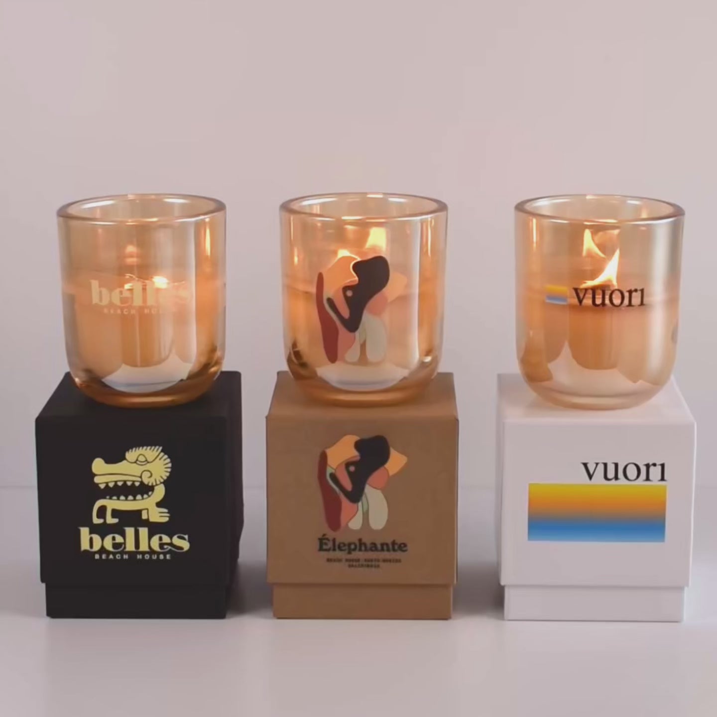 Load video: Three lit 12 oz Luxe Custom Candles in the color “Daybreak” which is a glimmering gold from Custom Candles LA. The 1st candle has a yellow &quot;Belles Beach House&quot; logo printed on the glass and sits on a black box that has the logo printed in yellow. The second candle has a multicolor &quot;Elephante&quot; logo on the glass and sits on top of a Kraft box with the same logo printed on it. The third candle has the &quot;Vuori&quot; logo printed on the glass in multicolor, and sits on a white box with the logo printed on it.