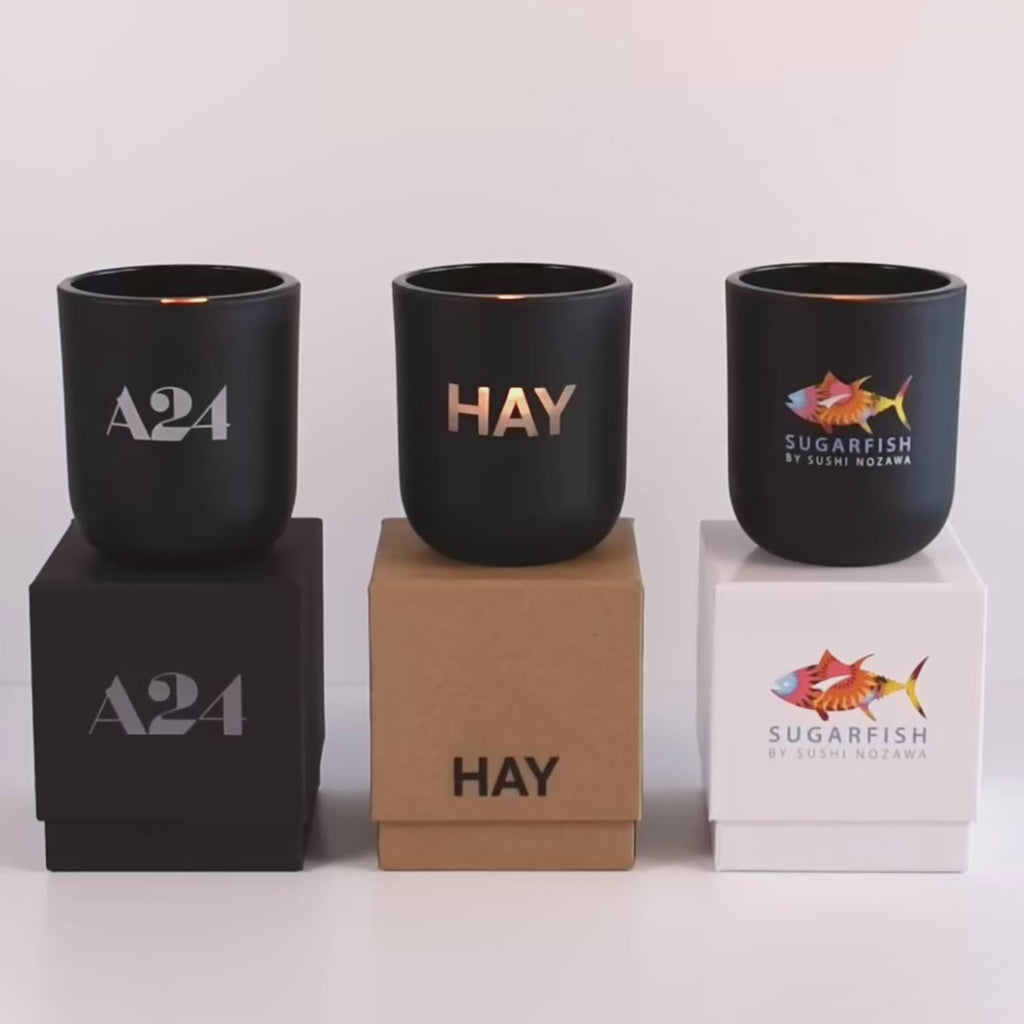 Three lit 12 oz Luxe Custom Candles in the color “Darkest Night” which is a jet black from Custom Candles LA. The first candle has a grey "A24" logo printed on the glass and sits on a black box that has the same logo. The second candle has an etched "HAY" logo on the glass and sits on top of a Kraft box with the same logo printed on it in black. The third candle has the multicolor "sugarfish by sushi nozawa" logo printed on the glass, and sits on a white box with the logo printed in multicolor.